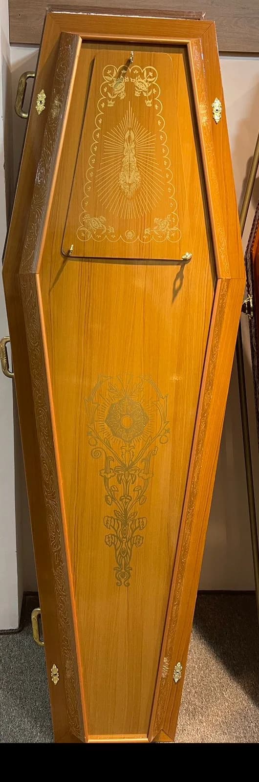 Urna funerária em madeira clara com gravura devocional e detalhes dourados.