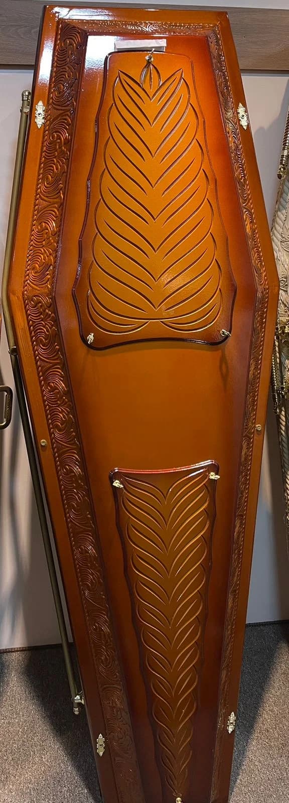 Urna funerária em tom mel com entalhe ornamental no painel frontal.